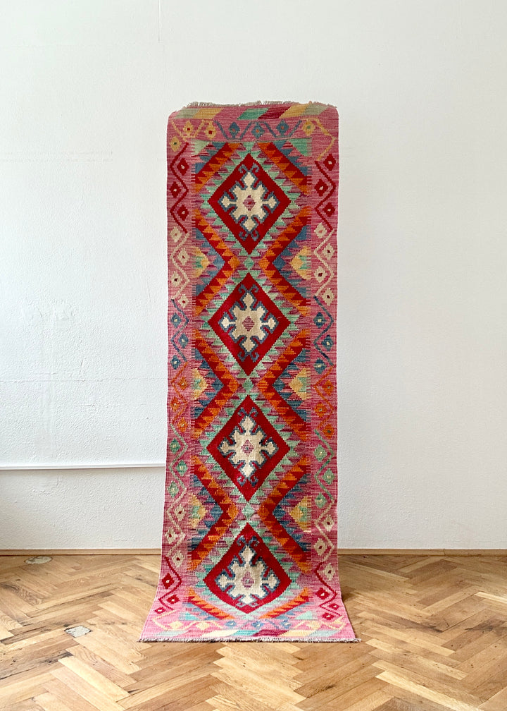 Frontalaufnahme eines pinkfarbenen Kelim-Läufers mit roten Rautenmedaillons und cremeweißen Sternmotiven 