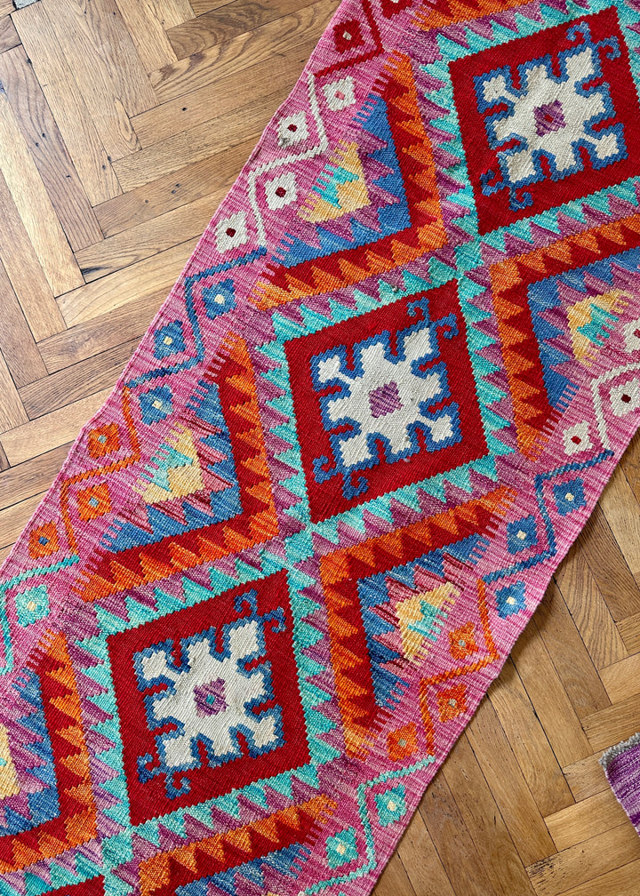 Detailaufnahme eines pinkfarbenen Kelim-Läufers mit roten Rautenmedaillons und cremeweißen Sternmotiven auf Parkettboden